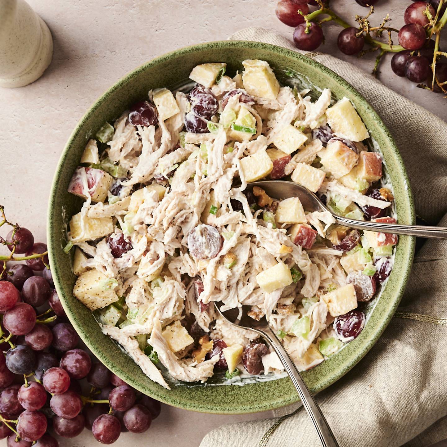 Overhead view of Waldorf chicken salad in a green bowl, made with shredded chicken, red grapes, celery, apples, and walnuts, served with two spoons.