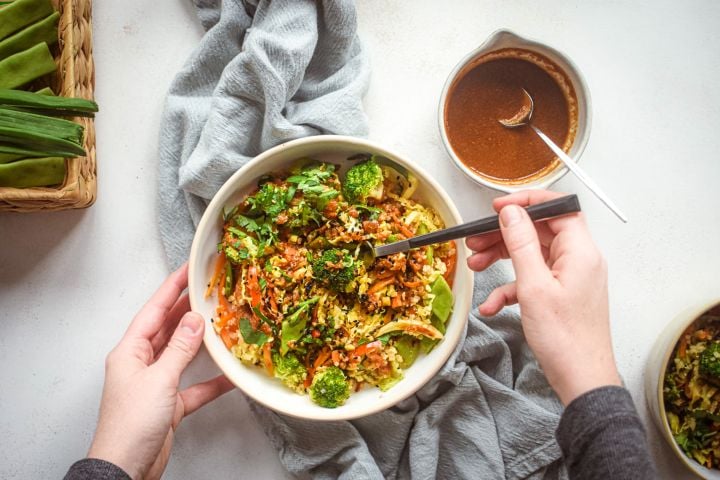 Asian quinoa stir fry with broccoli, carrots, cabbage, and snow peas in a bowl with stir fry sauce and sesame seeds.