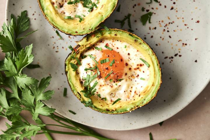 Two avocado halves baked with eggs, sprinkled with red pepper flakes and fresh chopped parsley, served on a neutral plate with parsley garnish on the side.