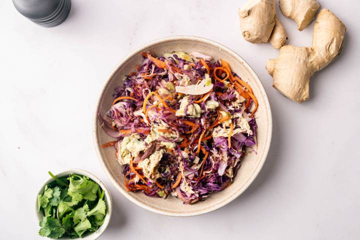A colorful salad with shredded purple cabbage, carrots, and dressing in a beige bowl on a white surface. Fresh cilantro and ginger root surround it, suggesting fresh ingredients.