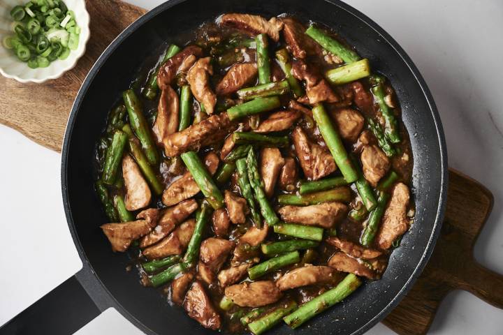 Stir-fried chicken and asparagus in a dark sauce, garnished with chopped green onions in a bowl on the side.