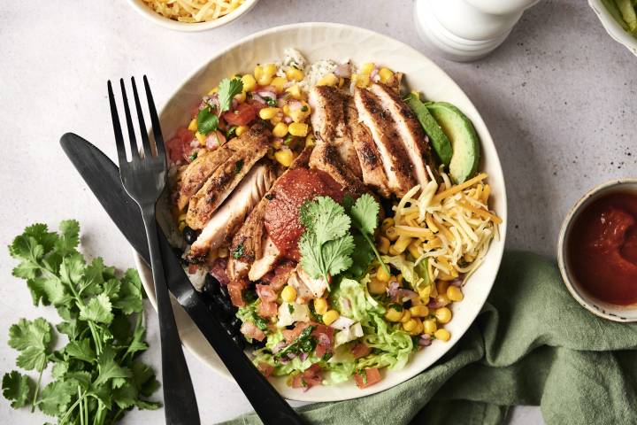 A colorful bowl featuring grilled chicken slices, corn, cheese, lettuce, and salsa, garnished with cilantro and avocado.