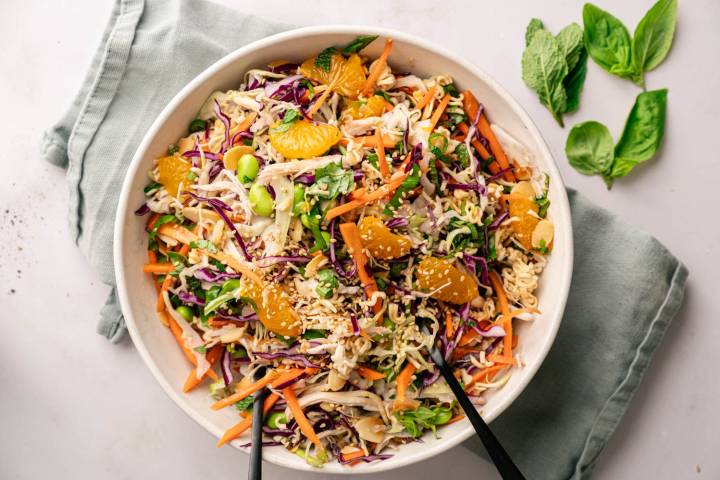 A vibrant salad in a white bowl features shredded cabbage, carrots, edamame, mandarin slices, and sesame seeds, on a pale green napkin with basil leaves nearby.