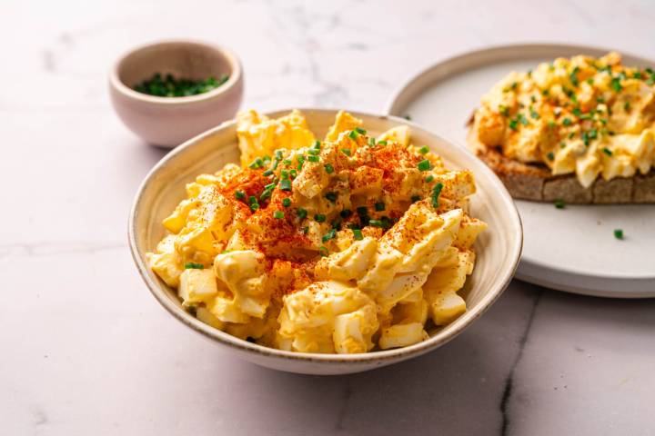 Bowl of egg salad topped with paprika and chopped chives, next to a slice of bread with egg salad. Small bowl of chives in the background.