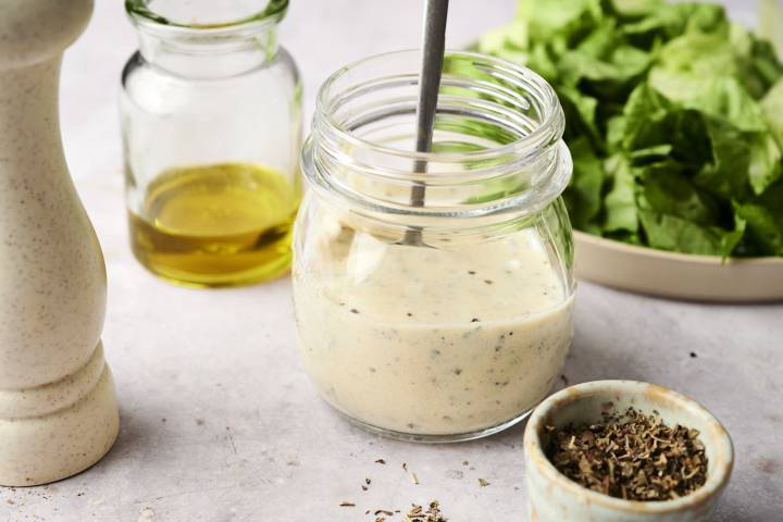 Jar of creamy Greek salad dressing with herbs, olive oil, and fresh lettuce in the background.
