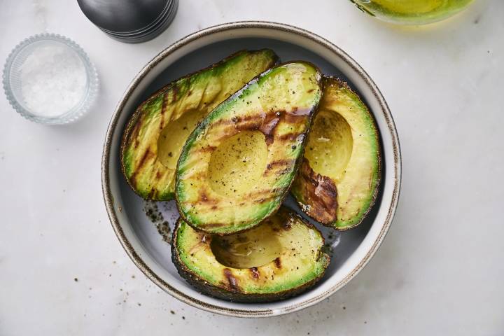 Grilled avocado halves drizzled with oil and sprinkled with pepper, served in a bowl beside coarse salt.