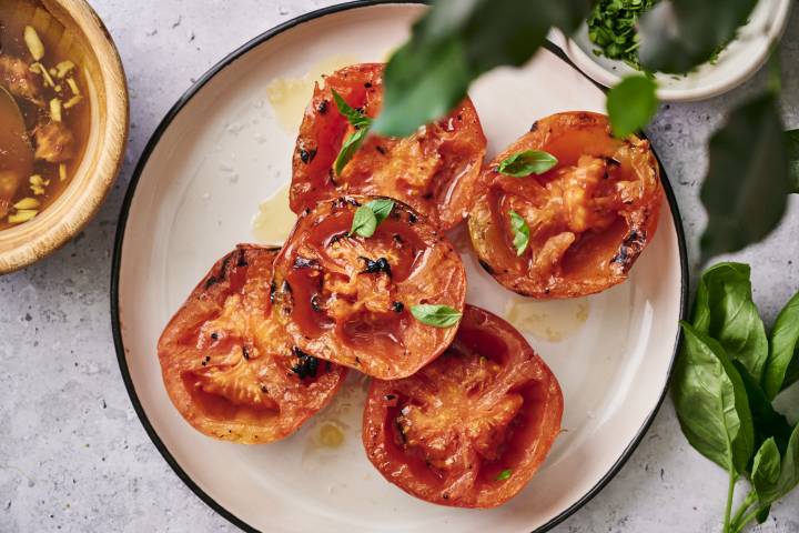 Grilled tomatoes topped with fresh basil, drizzled with olive oil, served on a round plate alongside a bowl of broth.