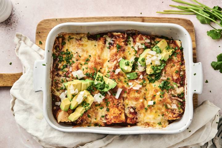 A savory enchilada filled with beans and chicken, topped with avocado, onion, and cilantro sits on a white plate.