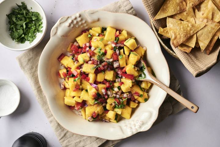Fresh mango salsa in a cream-colored bowl with a wooden spoon, made with diced mango, red onion, red bell pepper, jalapeño, and cilantro, served with tortilla chips on the side.