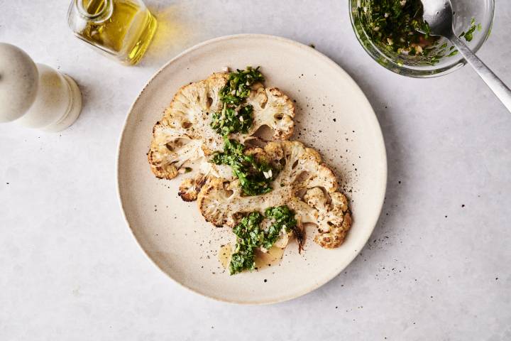 Overhead view of roasted cauliflower steaks garnished with chimichurri sauce, served on a plate with olive oil and pepper mill nearby.