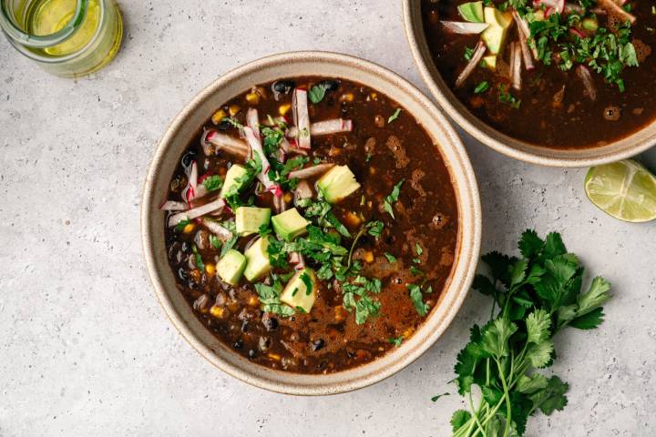 A bowl of black bean soup topped with avocado, cilantro, and radish strips on a textured gray surface. A jar of oil and fresh lime and cilantro are nearby.