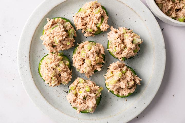 Six cucumber slices topped with tuna salad, garnished with celery and pepper, arranged on a white plate. The tone is fresh and appetizing.