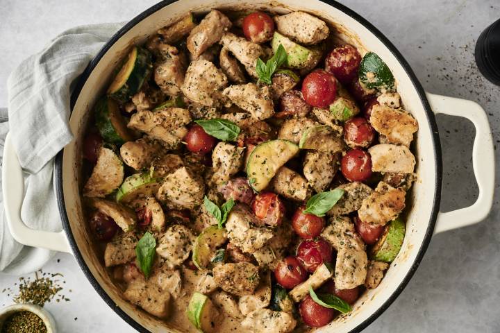 Top-down view of a creamy Tuscan chicken skillet filled with chunks of seasoned chicken, cherry tomatoes, zucchini slices, and fresh basil in a creamy herb sauce. A gray napkin and small bowl of Italian seasoning rest nearby.