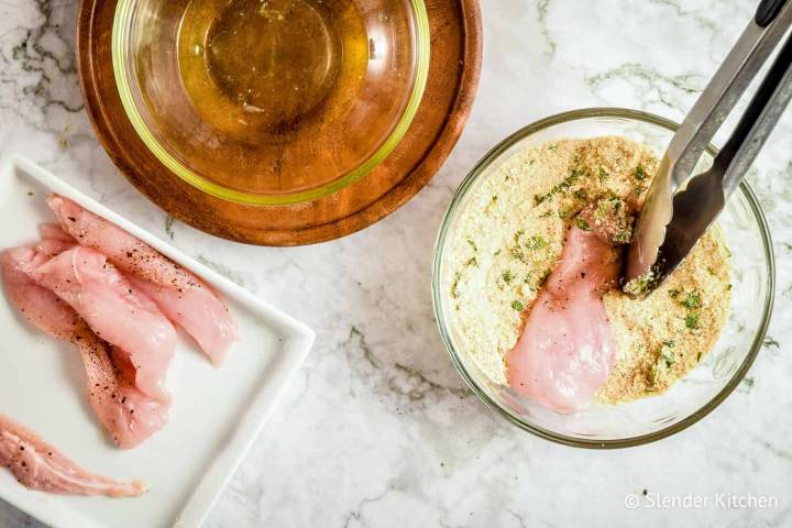 Chicken breast being dipped into Parmesan breadcrumbs with tongs.