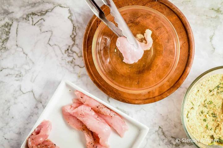 Chicken breast being dipped into egg whites with tongs.