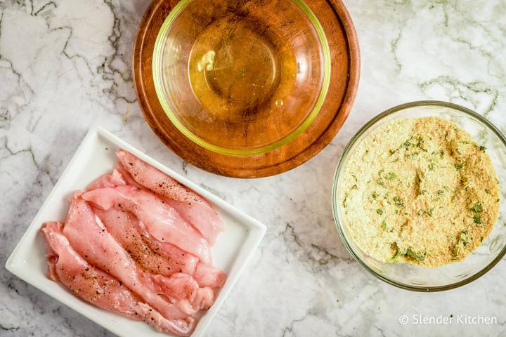 Plate with raw chicken breast, bowl with egg whites, and bowl with parmesan breading.