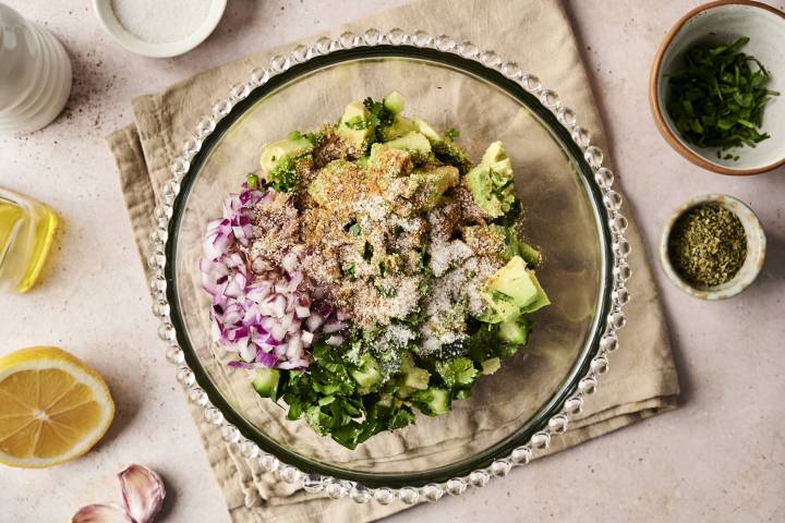 Glass bowl filled with diced avocado, cucumber, red onion, fresh cilantro, and a blend of seasonings, ready to be mixed into avocado cucumber salsa, surrounded by lemon, garlic, oil, and herbs