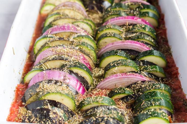 Herbs sprinkled on sliced eggplant, zucchini, and onion in a baking dish with crushed tomatoes.