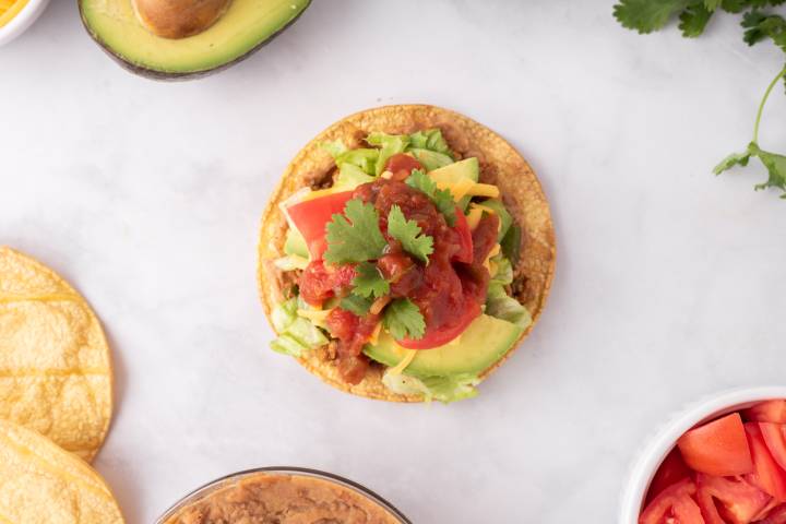Lettuce, avocado, tomato, cilantro, and salsa being added to a tostada. 