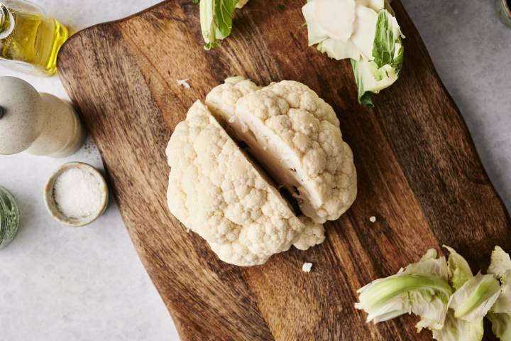 Whole cauliflower partially sliced in half on a wooden cutting board with seasoning ingredients around.