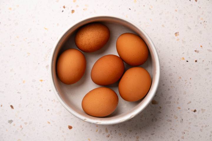 A white bowl containing six brown eggs is placed on a speckled countertop.
