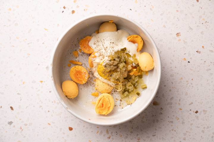 A white bowl on a speckled surface contains sliced hard-boiled egg yolks, creamy sauce, chopped pickles, and a sprinkle of black pepper.