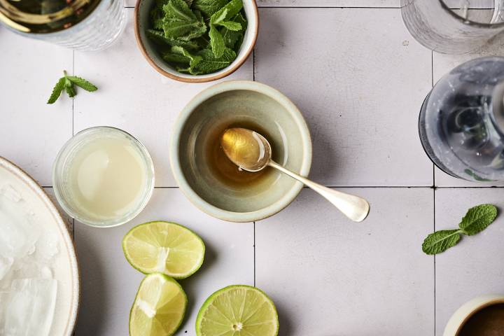 A flat lay of fresh mint, lime halves, syrup in a bowl, and a glass of lime juice on a light tiled surface.