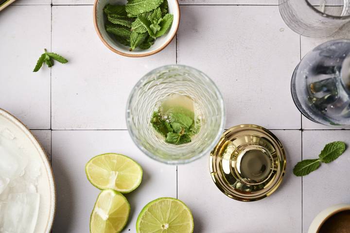 A top-down view of a cocktail glass filled with mint and lime, surrounded by lime halves, mint leaves, and a bowl of mint.