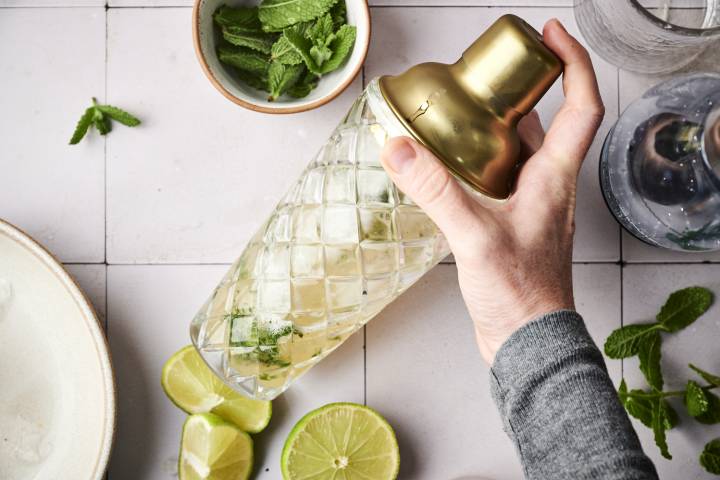 A hand shakes a cocktail shaker filled with mint, lime, and ice, surrounded by fresh mint leaves and lime slices on a tiled surface.