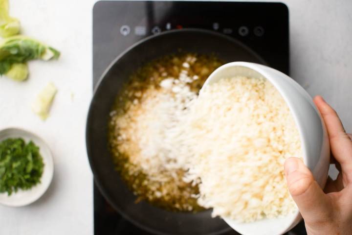 Cauliflower rice being added to a pan with broth, onions, and garlic.