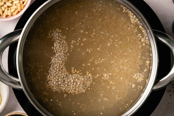 Pot of quinoa simmering in water, forming gentle bubbles on the surface. A bowl of pine nuts is partially visible, suggesting a cooking process.