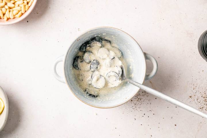 A white bowl filled with creamy pasta and black truffle slices, with a spoon resting inside. Nearby are a dish of pine nuts and a lemon slice, creating a gourmet cooking scene.