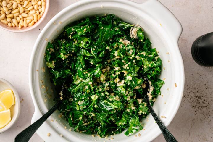 A white bowl filled with a vibrant kale and quinoa salad, topped with crumbled cheese. Nearby are bowls of pine nuts and lemon wedges, setting a fresh, healthy tone.