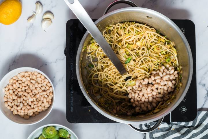 White beans being added to a pot of pasta and shredded Brussels sprouts.