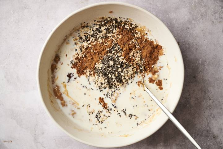 A top-down shot of a person pouring milk into a jar with oats, chia seeds, and banana slices.