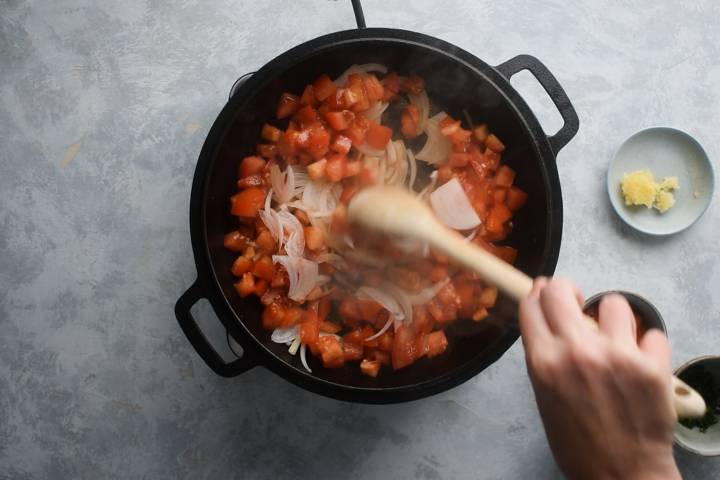 Tomatoes and onions cooking in a cast iron skillet with a wooden spoon.