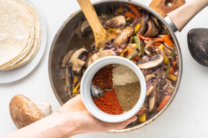 Fajita seasoning being added to a skillet with peppers, onion, and portobellos.