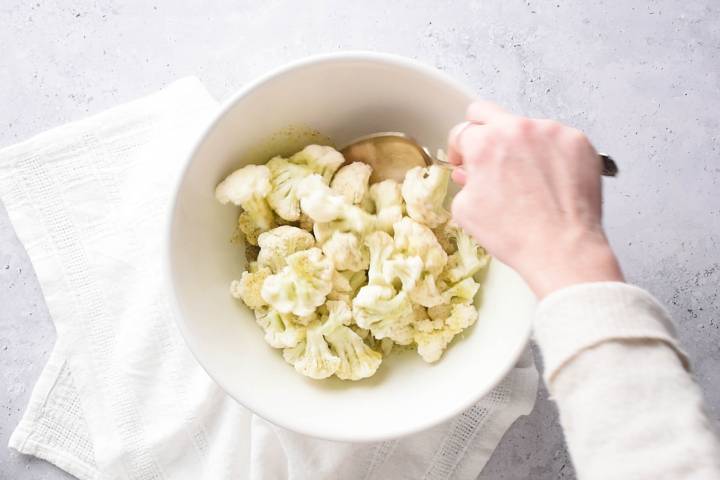 Cauliflower florets in a bowl being tossed with olive oil, garlic powder, salt, and pepper.