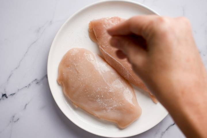 Chicken breast being seasoned with salt and pepper.