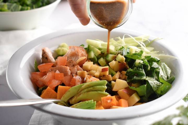 Salmon, kale, avocado, and sweet potato salad with dressing being drizzled on top.