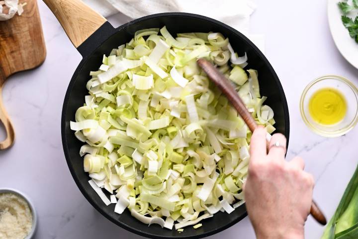 Leeks in a large skillet being stirred with a wooden spoon.