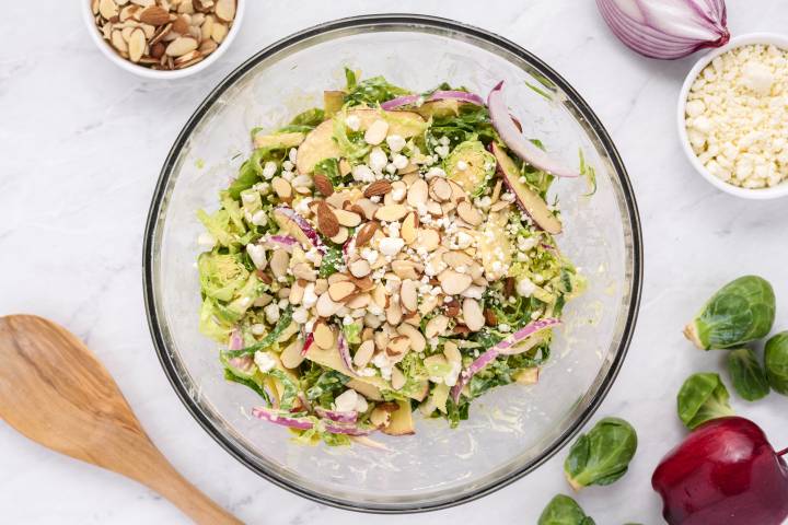 Shaved brussels sprouts salad in a glass bowl with apples, goat cheese, and almonds.