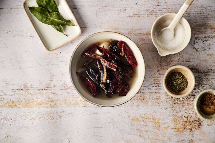 Dried chili peppers are soaking in a bowl of water, surrounded by bay leaves, salt, and spices, preparing the base for Mexican beef adobo.