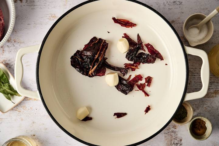 Dried chili peppers and garlic cloves are toasting in a white pot, enhancing their flavors before blending into the adobo sauce.