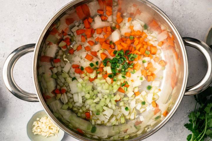Chopped onions, carrots, celery, and green peppers in a silver pot with olive oil. A small dish of minced garlic is beside the pot. Fresh herbs are nearby. 