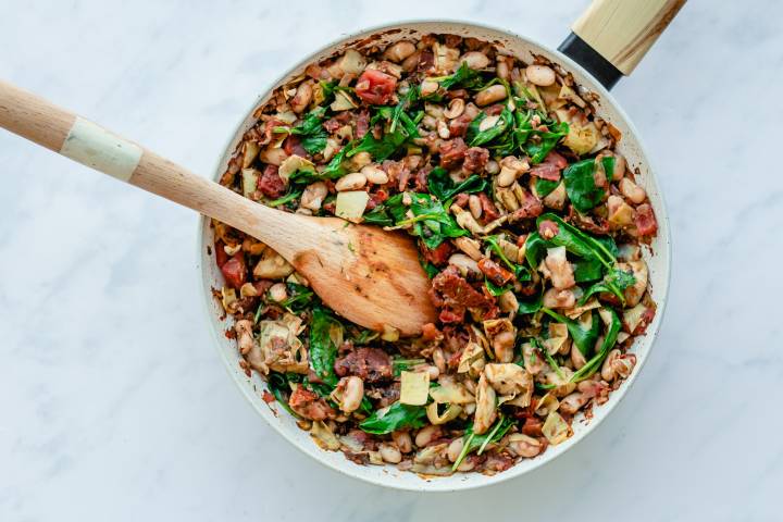 Spinach being added to tomatoes, white beans, and artichokes in a skillet.