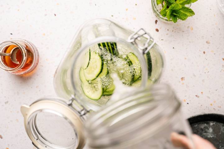 Top-down view of a glass jar filled with water and cucumber slices. Nearby is a jar of honey and fresh mint leaves, on a speckled surface. 