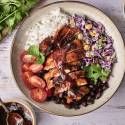 A colorful bowl featuring grilled chicken, rice, black beans, cherry tomatoes, slaw, and herbs, drizzled with barbecue sauce.