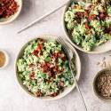 Two bowls of creamy broccoli salad topped with bacon and seeds are on a marble surface. Nearby are small bowls of bacon bits, sunflower seeds, and a fork.