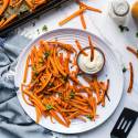 Crispy baked carrot fries garnished with fresh parsley on a white plate, served with a small bowl of creamy dipping sauce. The plate is placed on a marble countertop, surrounded by a baking tray, a blue fringed napkin, and scattered carrot fries and parsley.